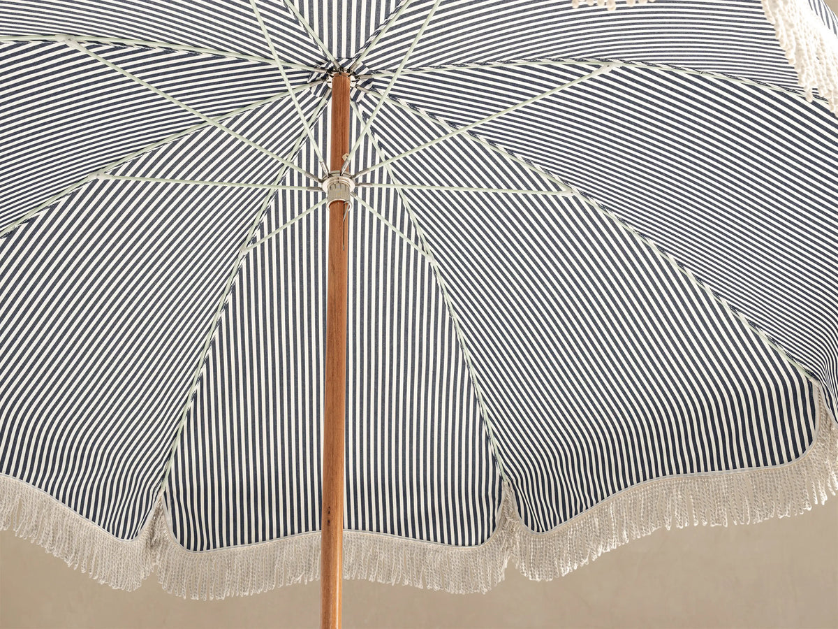 Front view of a navy and white striped outdoor umbrella with white fringe, a wooden pole, and a white scalloped base.