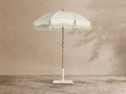 Front view of a cream patio umbrella with fringe trim, a wooden pole, and a white square base.