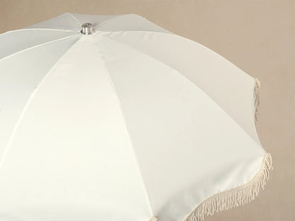 Front view of a cream patio umbrella with fringe trim, a wooden pole, and a white square base.
