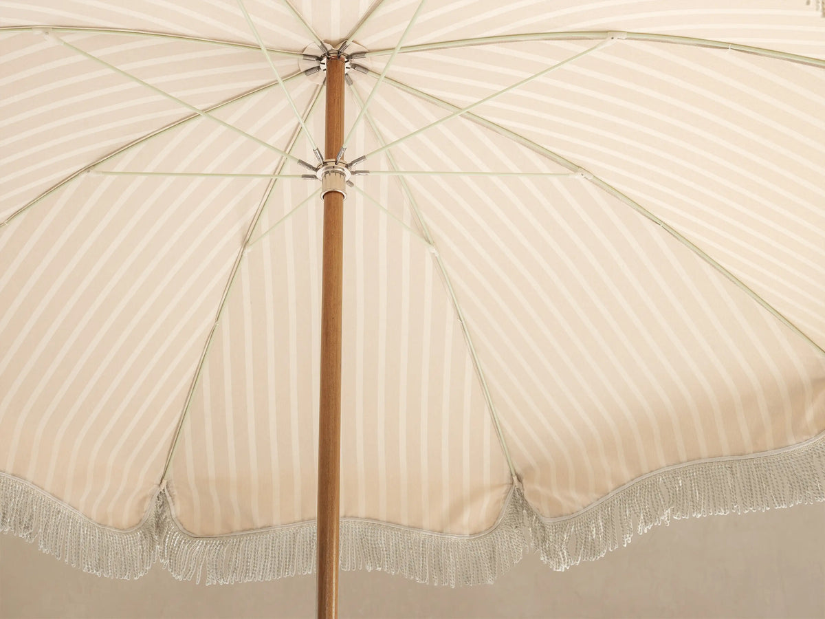 Underside view of a beige and white striped parasol with a wooden pole, metal spokes, and a light-colored fringe border.