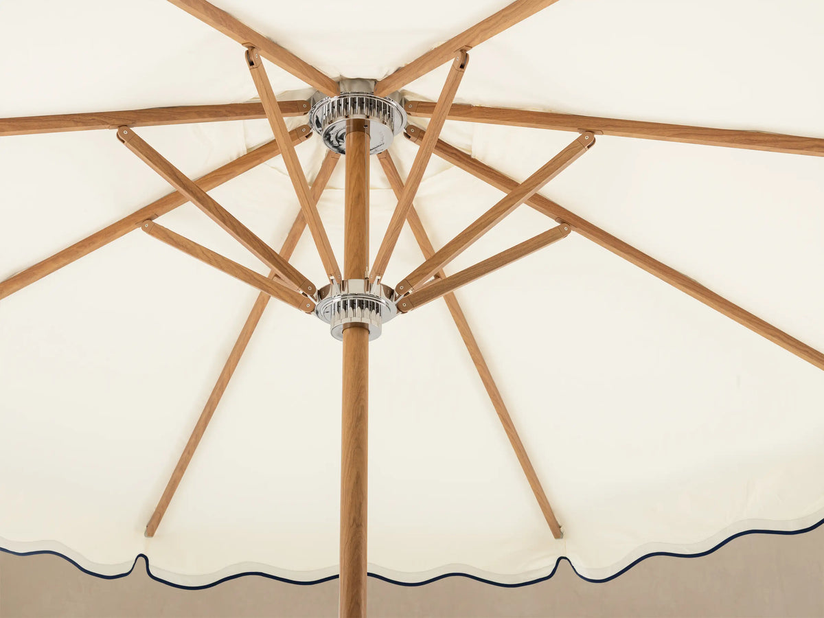 Front view of an off-white umbrella with a scalloped edge and a navy blue wavy stripe, on a light brown wooden pole and white base.