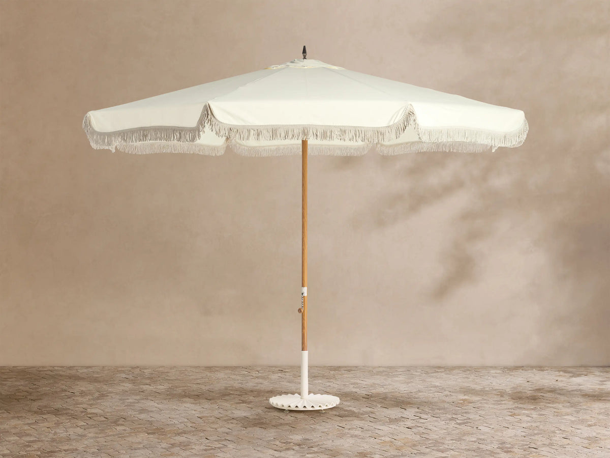 Front view of a cream patio umbrella with decorative fringe, a wooden pole, and a white base.