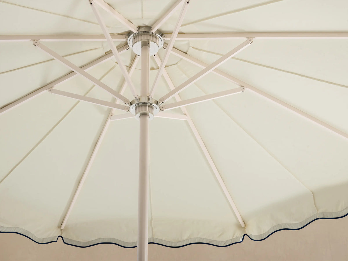 Front view of a cream patio umbrella with a scalloped edge and navy wavy trim on the canopy. White pole and base.