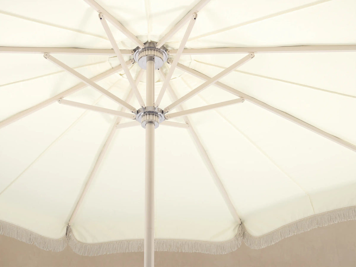 Front view of a Cream patio umbrella with a scalloped canopy edge and white fringe, on a white pole and round base.