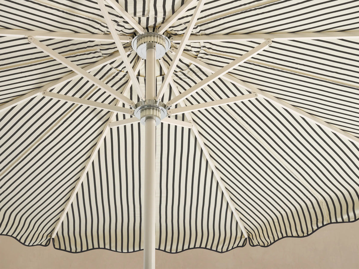 Front view of a cream and black vertically striped patio umbrella with a scalloped edge and a white pole and base.