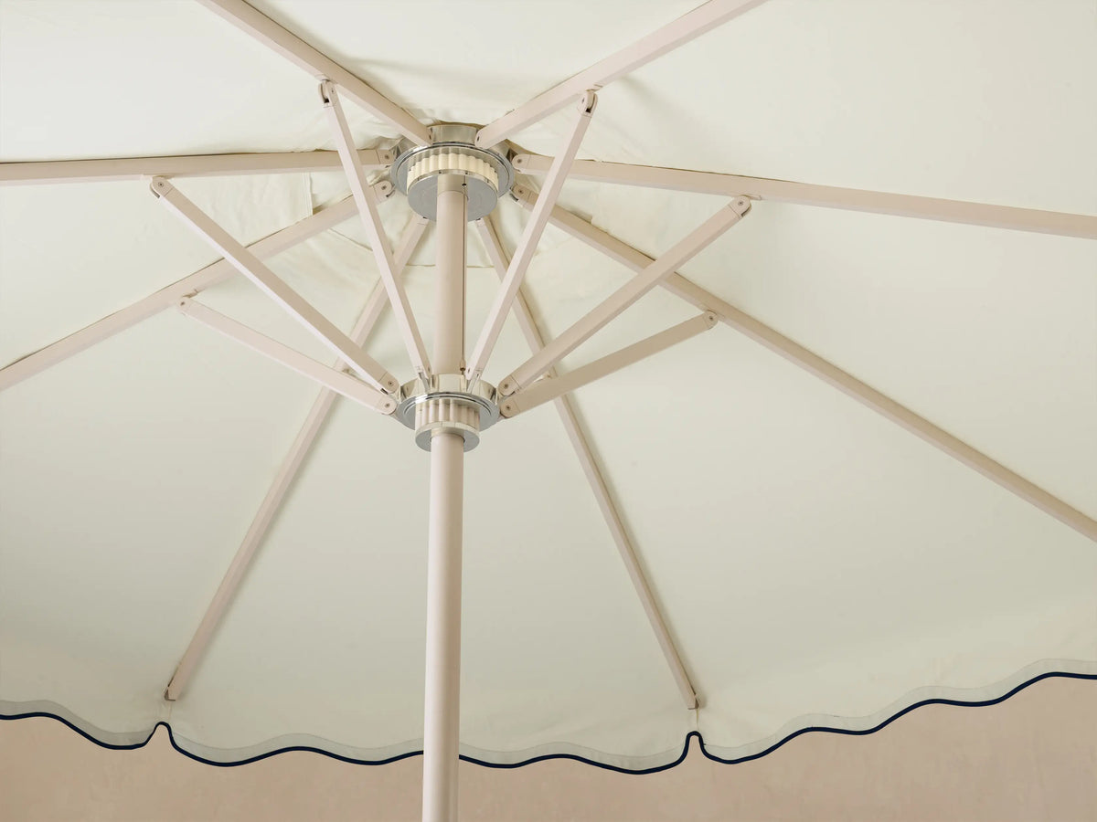 Interior view of an open cream umbrella with light tan ribs, a silver hub, and a scalloped edge with navy trim.