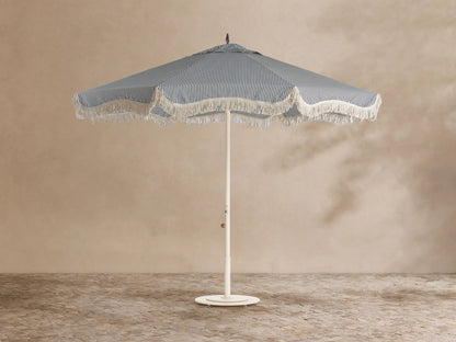 Front view of a blue and white vertical striped beach umbrella with scalloped edge and white fringe, on a cream pole.