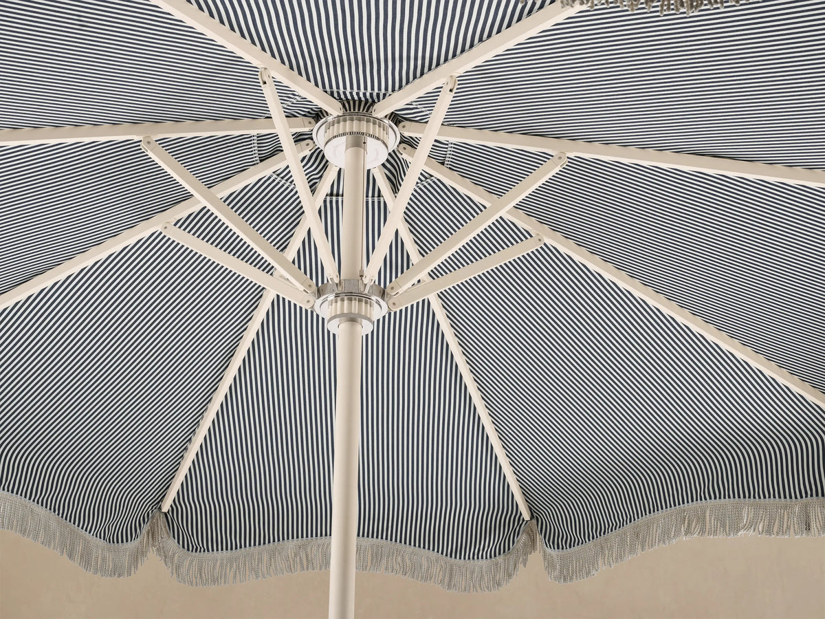 Front view of a blue and white vertical striped beach umbrella with scalloped edge and white fringe, on a cream pole.