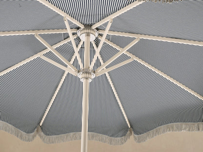 Front view of a blue and white vertical striped beach umbrella with scalloped edge and white fringe, on a cream pole.