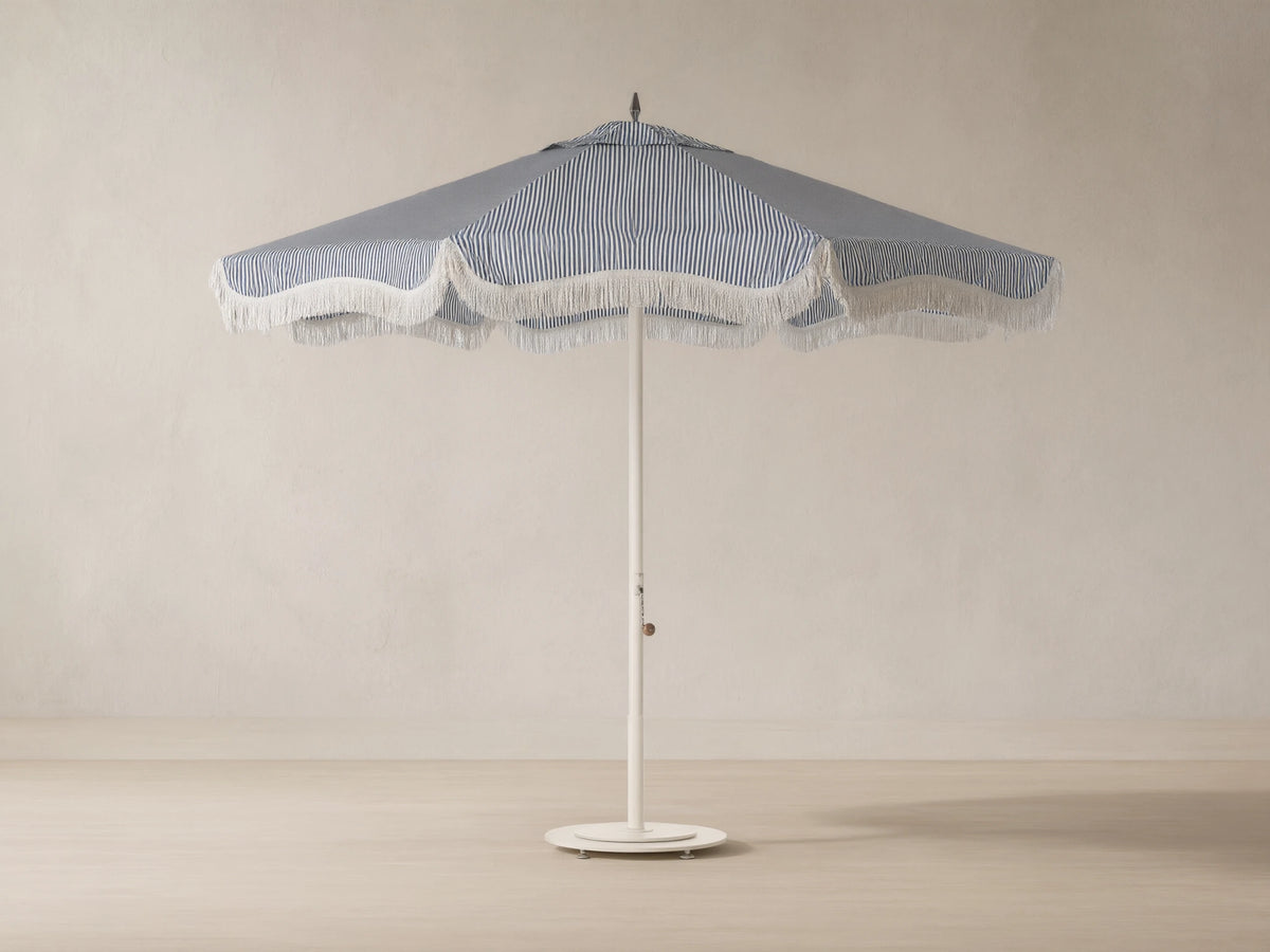 Front view of a blue and white vertical striped beach umbrella with scalloped edge and white fringe, on a cream pole.