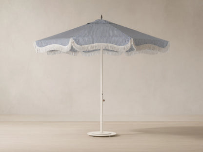 Front view of a blue and white vertical striped beach umbrella with scalloped edge and white fringe, on a cream pole.