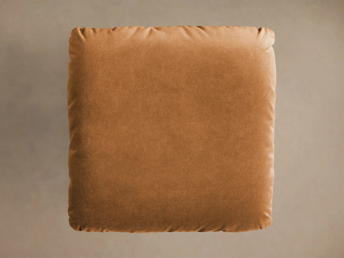 Front view of the caramel Deacon Ottoman, a plush square cushion on a shiny gold pedestal base.