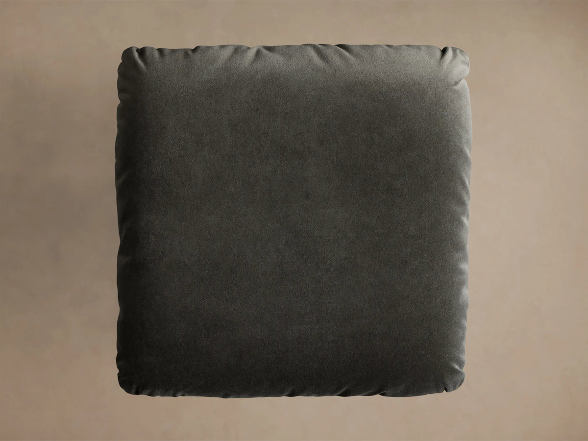 Front view of the charcoal Deacon Ottoman, featuring a plush cushion and a brushed metal pedestal base.