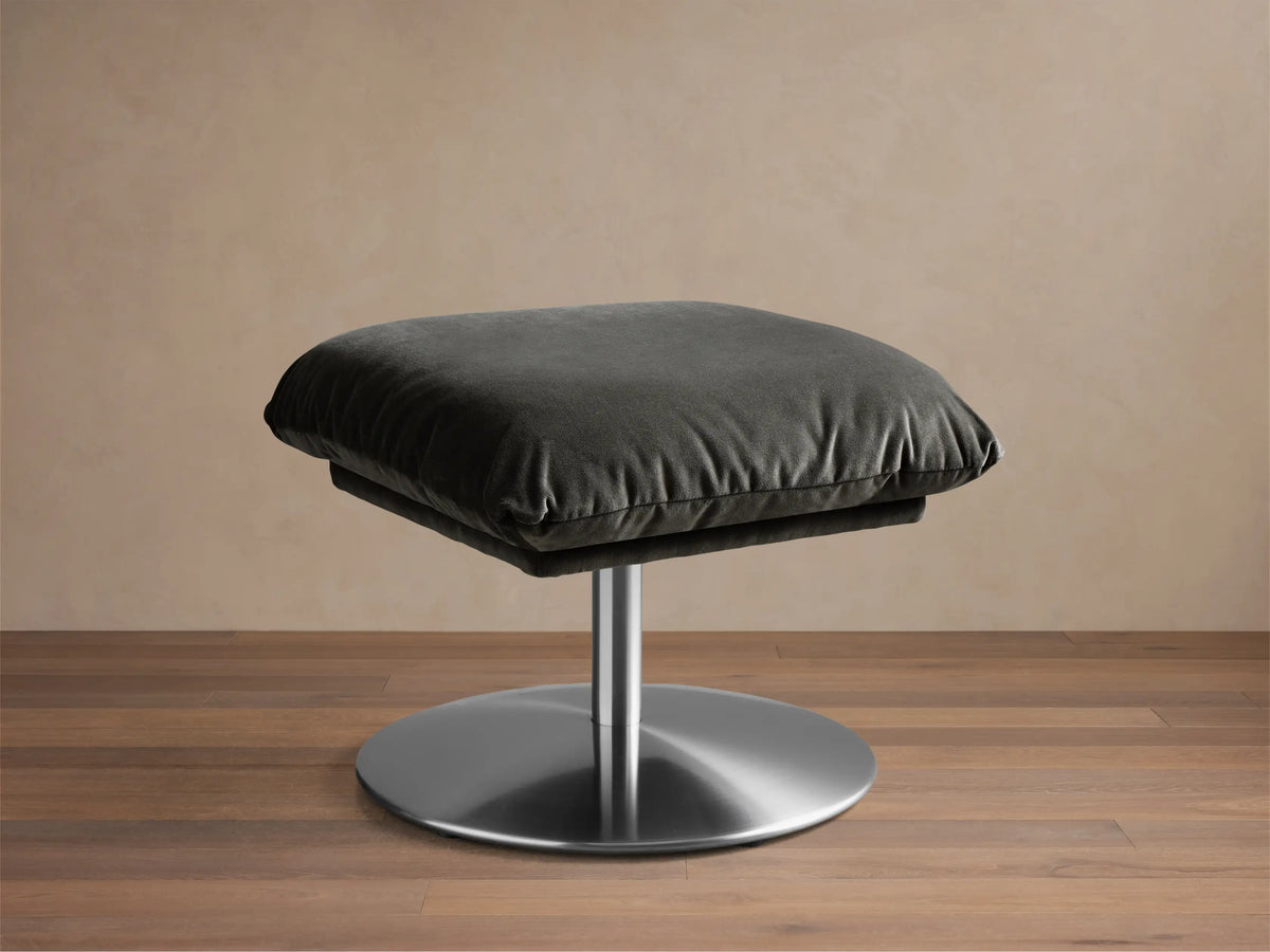 Close-up of the dark grey plush cushion of the Deacon Ottoman on its circular brushed metal pedestal base.