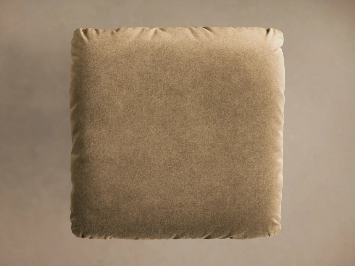 Close-up of the tan fabric cushion and brushed metal pedestal base of the Deacon Ottoman.