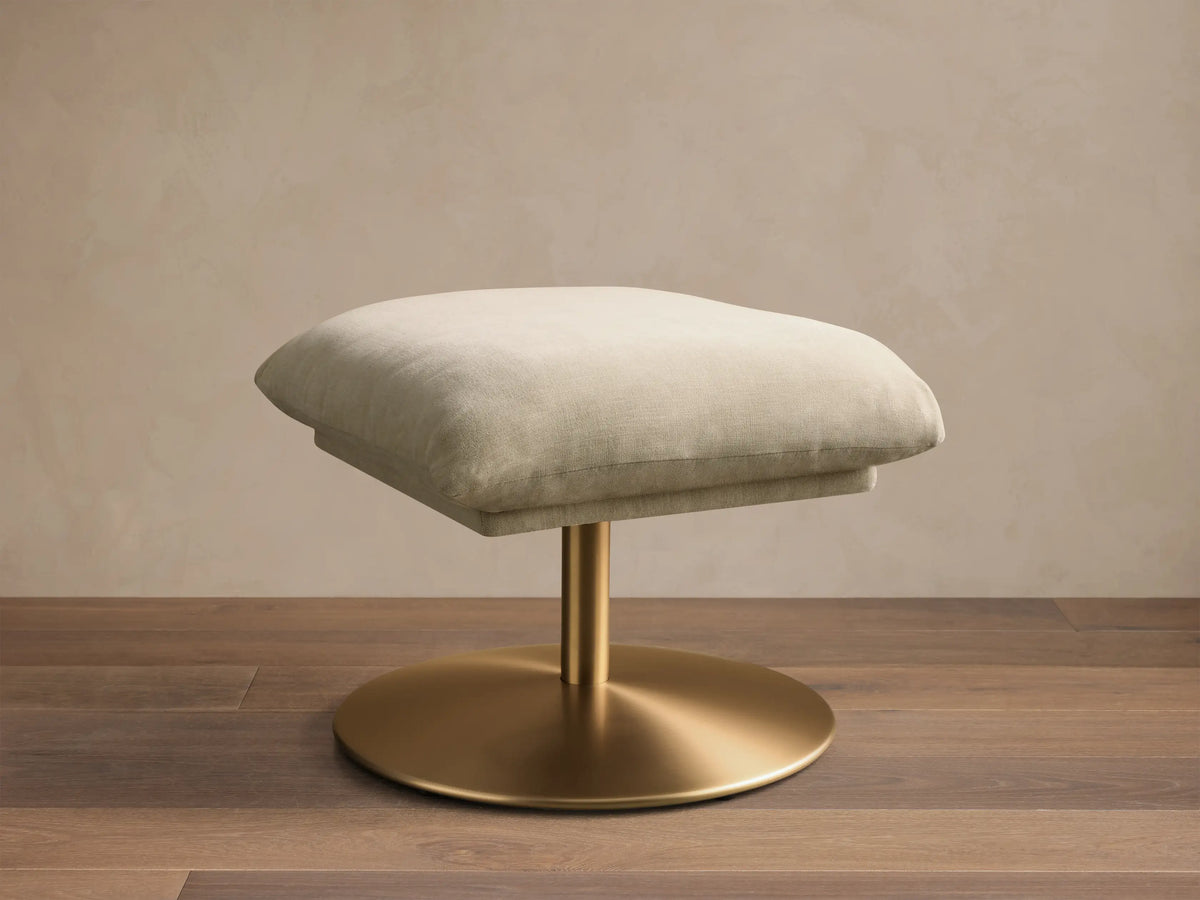 Front view of the cream Deacon Ottoman, featuring a soft padded cushion on a brushed gold pedestal base.