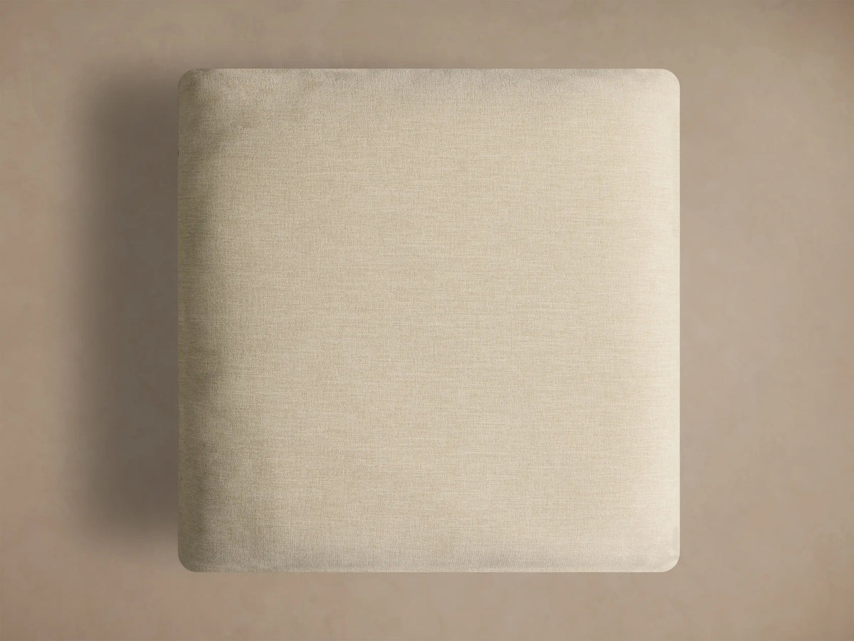 Close-up, angled view of the light beige textured cushion of the Deacon Ottoman and its golden metal base.