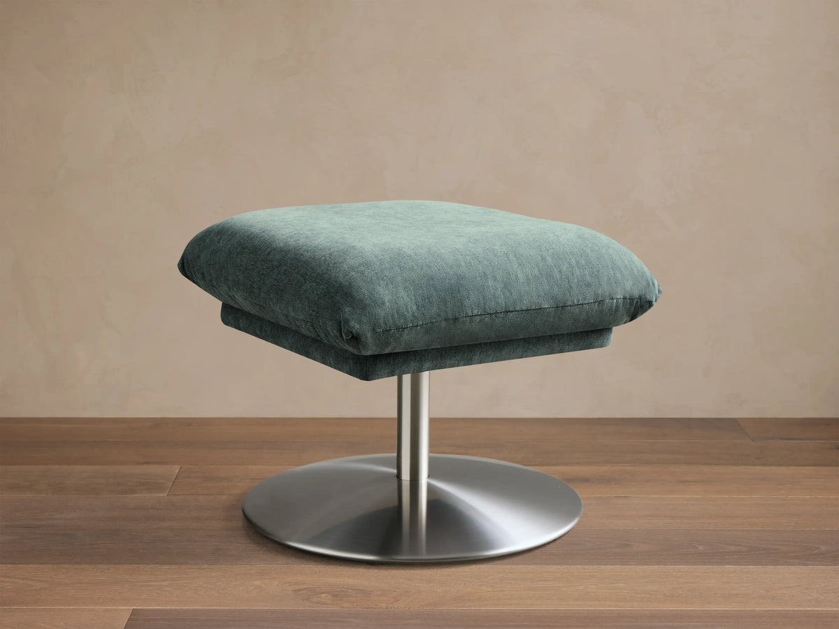 Front view of the teal upholstered Deacon Ottoman with a soft cushion and round silver metal base.