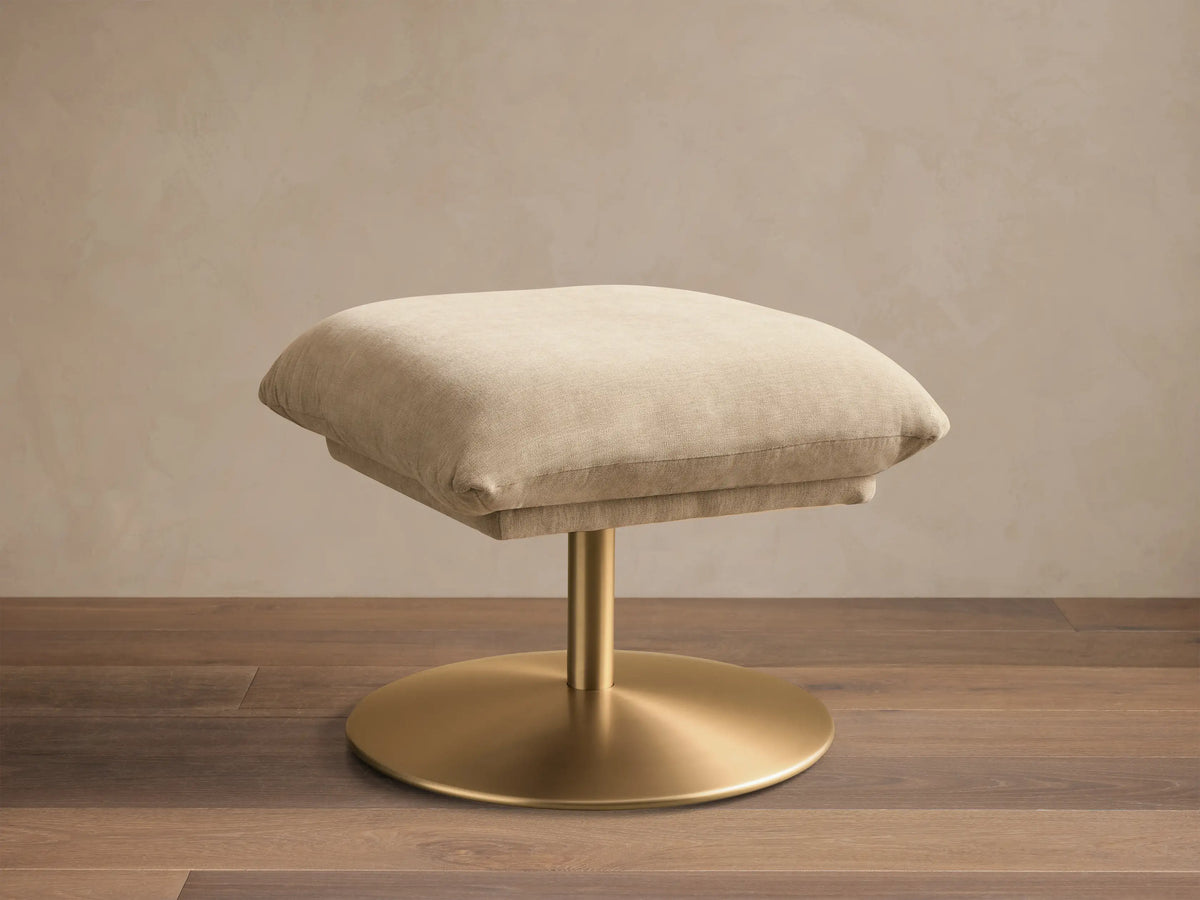 Front view of the beige Deacon Ottoman with a gold pedestal base.