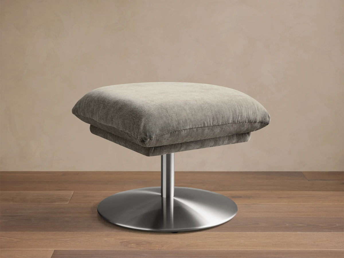 Front view of the Deacon Ottoman, featuring a light grey fabric cushion on a silver brushed metal pedestal base.