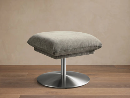 Front view of the Deacon Ottoman, featuring a light grey fabric cushion on a silver brushed metal pedestal base.