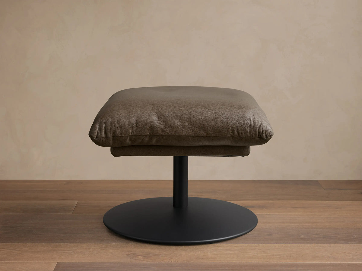 Front view of the taupe leather Deacon Leather Ottoman with a plump cushion and black circular pedestal base.