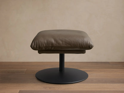Front view of the taupe leather Deacon Leather Ottoman with a plump cushion and black circular pedestal base.