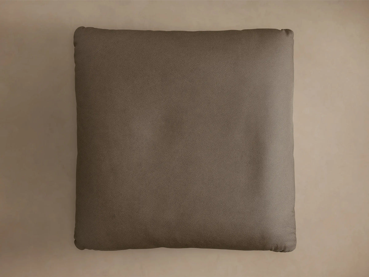 Front view of the taupe leather Deacon Leather Ottoman with a plump cushion and black circular pedestal base.