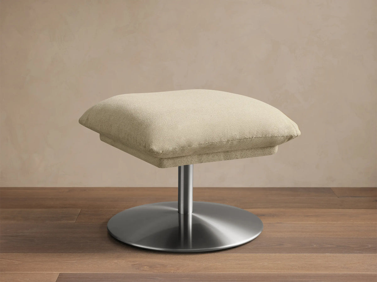 Front view of the cream Deacon Ottoman with a textured cushion and a silver-toned metal base.