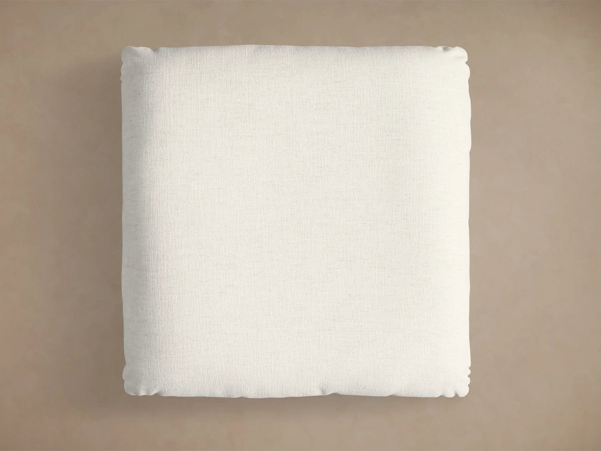 Close-up of the light beige textured cushion and circular brushed gold base of the Deacon Ottoman.