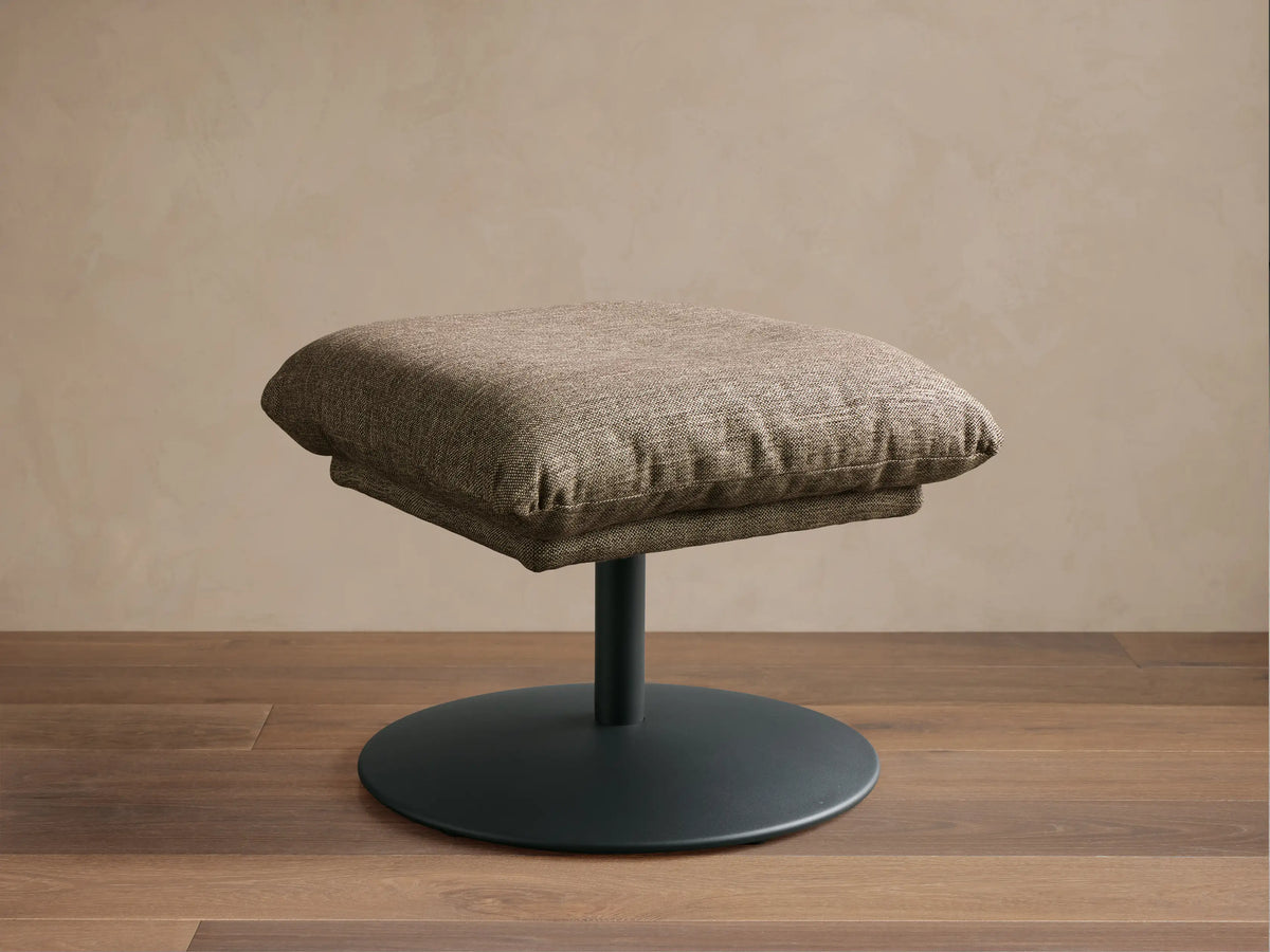 Front view of a textured light brown Deacon Ottoman with a plump square cushion on a sleek black round pedestal base.