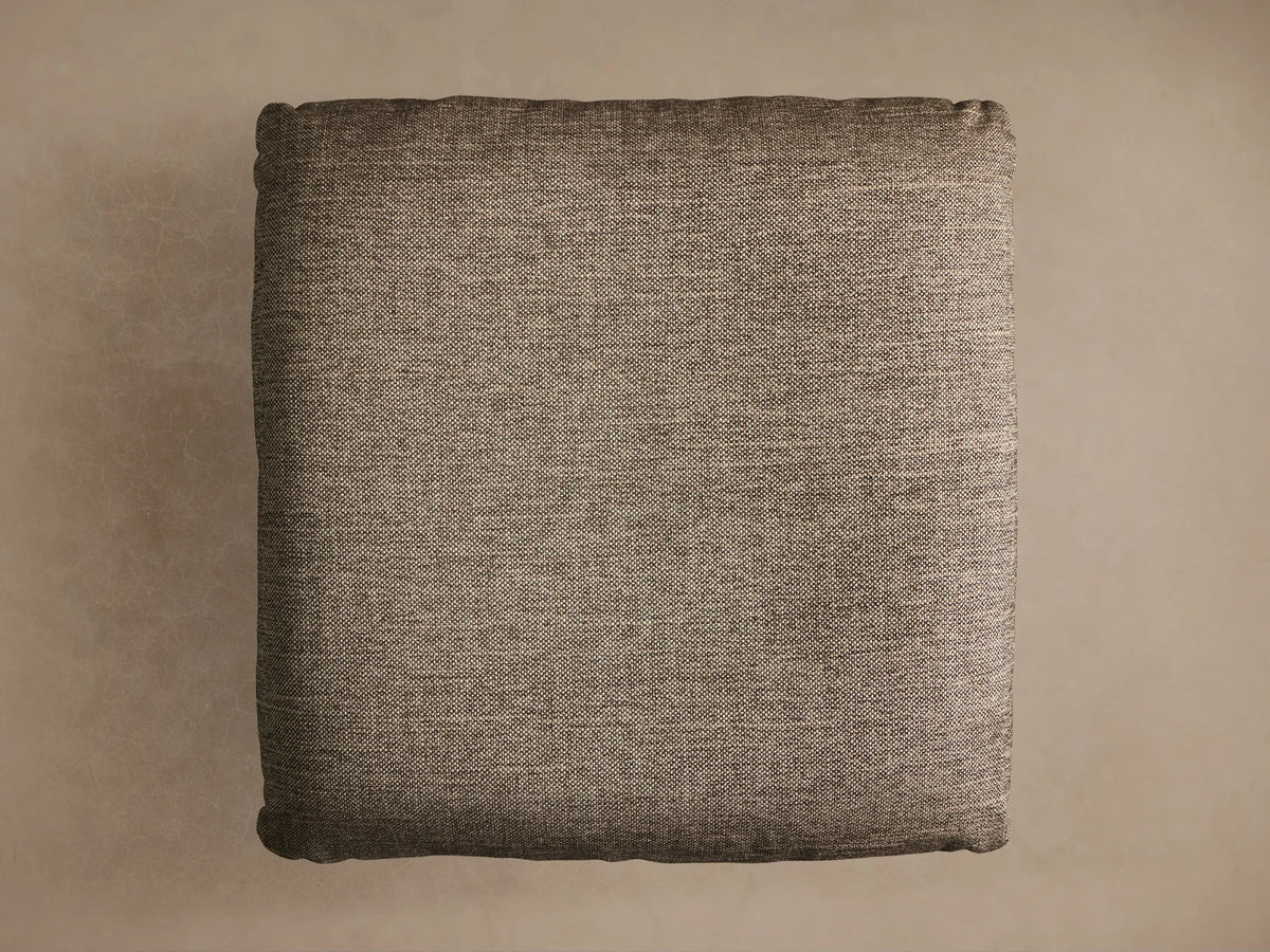 Close-up of the Deacon Ottoman. Angled view of its brownish-gray textured fabric cushion, atop a black circular pedestal base.