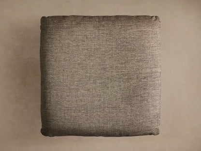 Close-up of the Deacon Ottoman. Angled view of its brownish-gray textured fabric cushion, atop a black circular pedestal base.