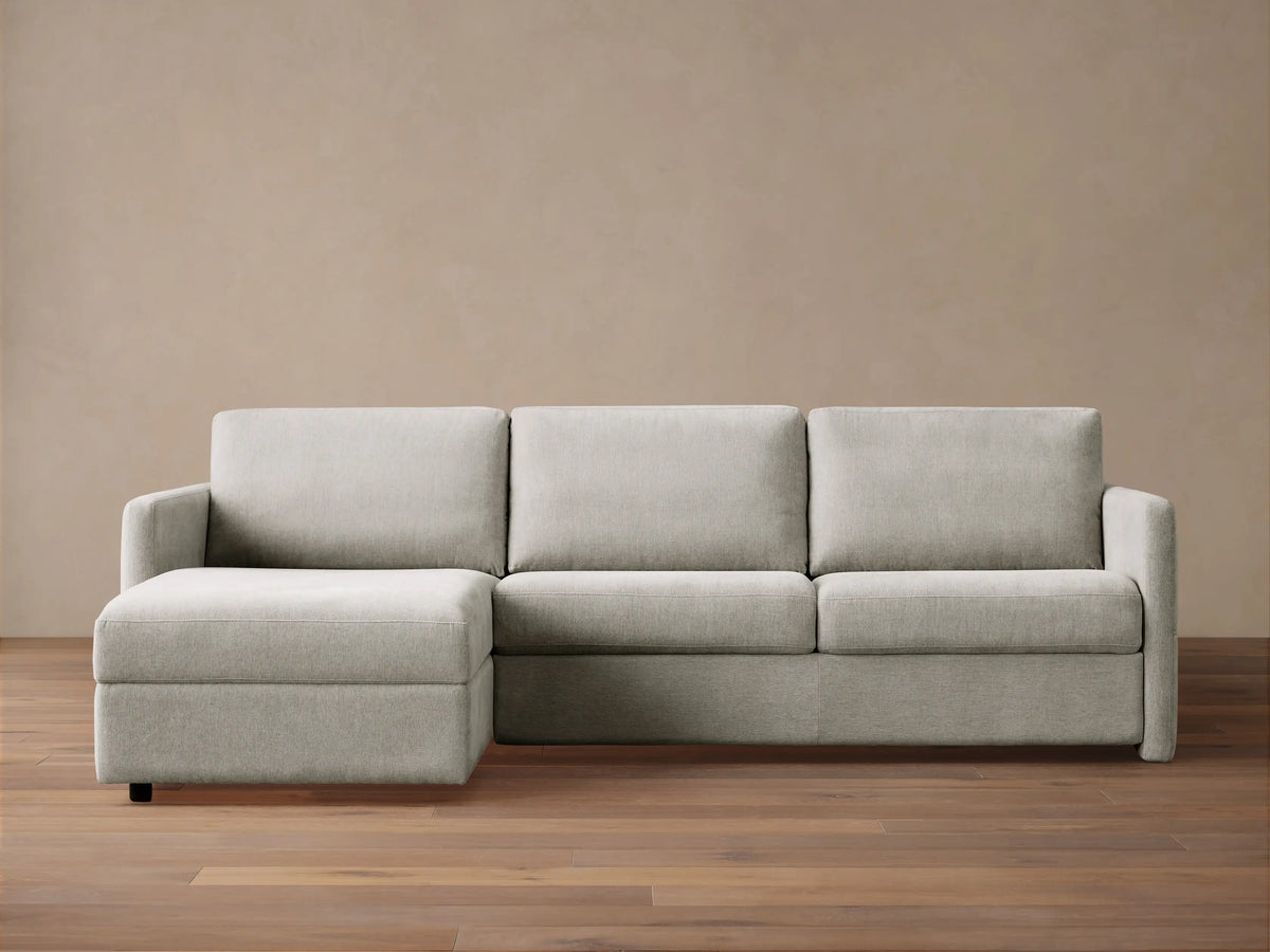 Light grey Reece Two-Piece Sleeper Chaise Sectional with a left-facing chaise, in a room with a light brown wall & wood floor.