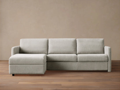 Light grey Reece Two-Piece Sleeper Chaise Sectional with a left-facing chaise, in a room with a light brown wall & wood floor.
