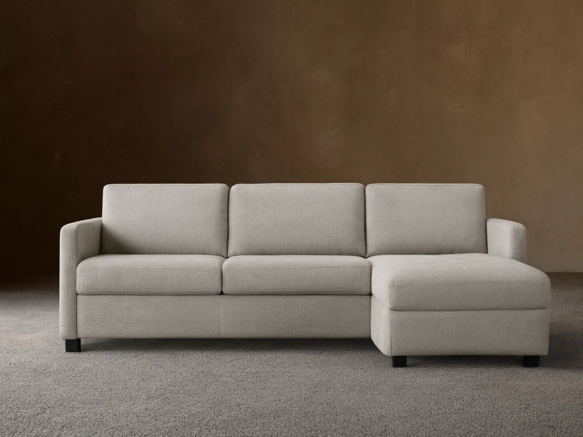 Light grey Reece Two-Piece Sleeper Chaise Sectional with a left-facing chaise, in a room with a light brown wall & wood floor.