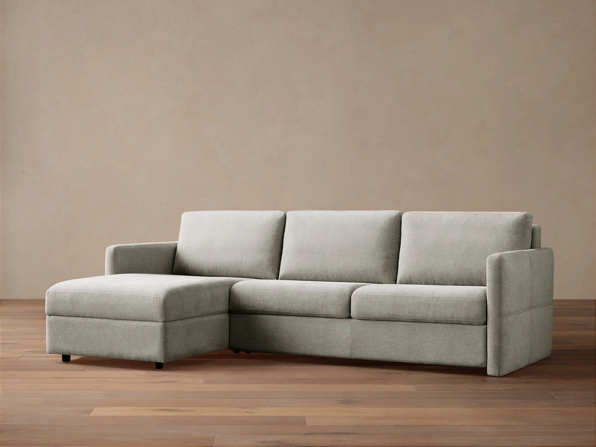 Light grey Reece Two-Piece Sleeper Chaise Sectional with a left-facing chaise, in a room with a light brown wall & wood floor.