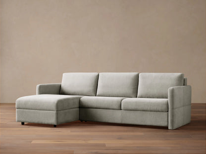 Light grey Reece Two-Piece Sleeper Chaise Sectional with a left-facing chaise, in a room with a light brown wall & wood floor.
