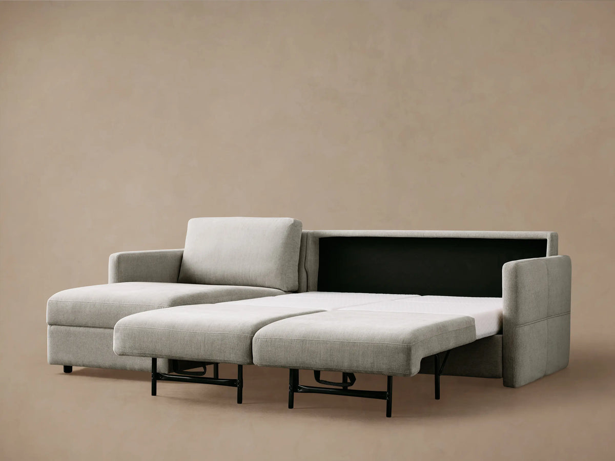 Light gray Reece Sleeper Chaise Sectional with its white, quilted foam mattress partially extended, against a dark background.