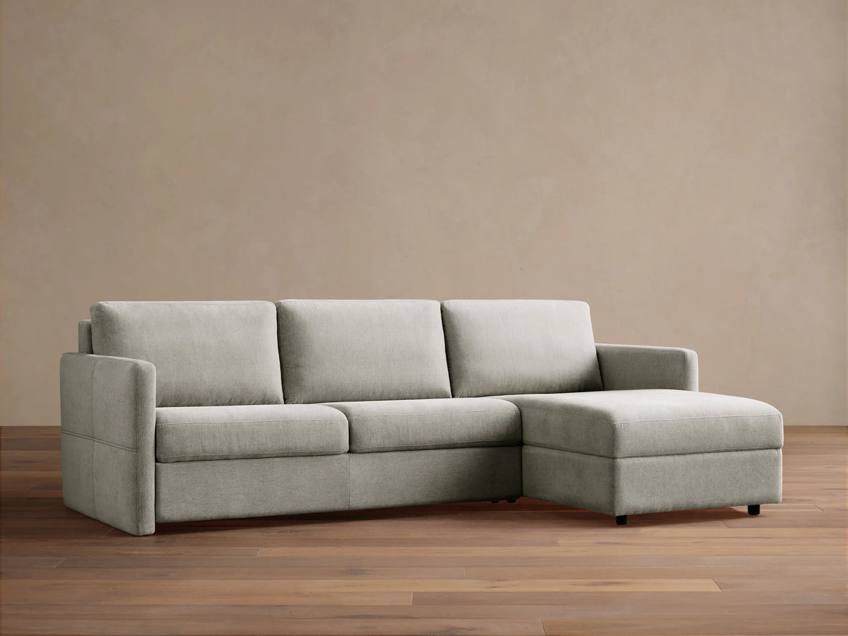 Light grey Reece Two-Piece Sleeper Chaise Sectional with an L-shape, chaise on the right, against a warm brown wall.