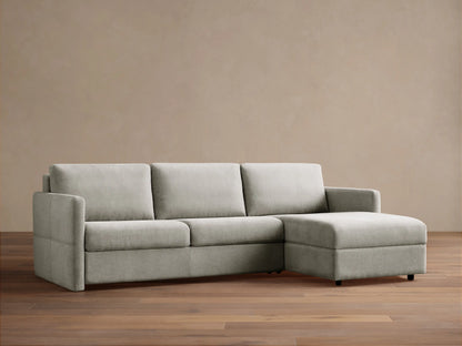 Light grey Reece Two-Piece Sleeper Chaise Sectional with an L-shape, chaise on the right, against a warm brown wall.