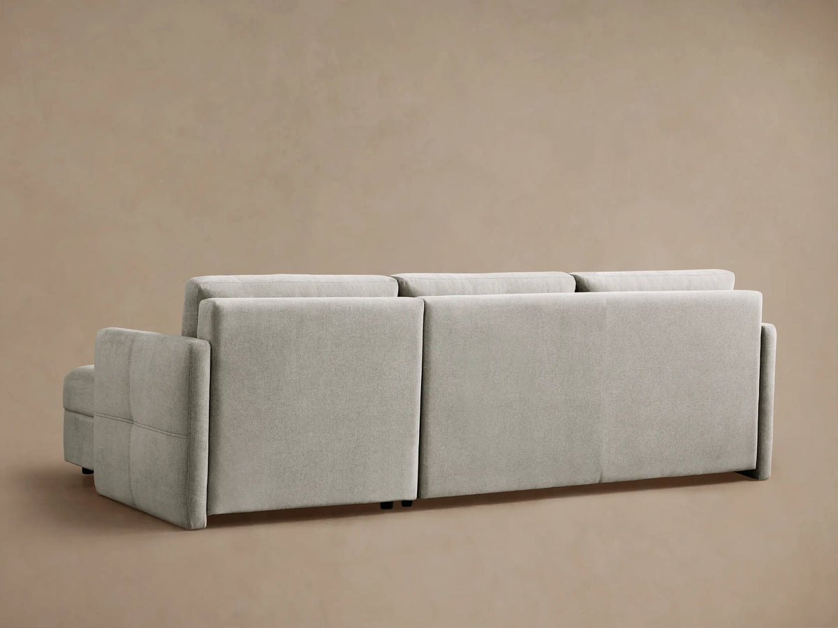 Light gray Reece Two-Piece Sleeper Chaise Sectional, left side converted to bed with white quilt & pillows, chaise right.