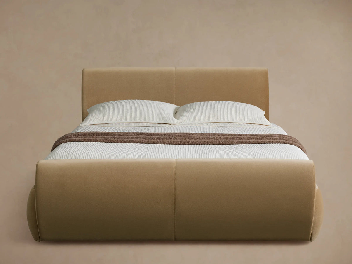 Blush pink upholstered Wynn Bed with a curved headboard and footboard, dressed in white bedding, in a bright bedroom.