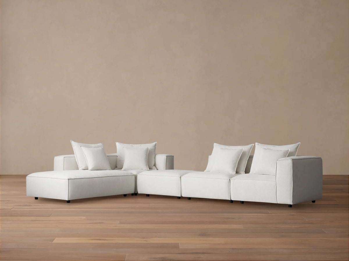Off-white Coburn Five Piece Bumper Sectional, L-shaped, with chaise, ottoman, and 3 armless seats, on wood floor.