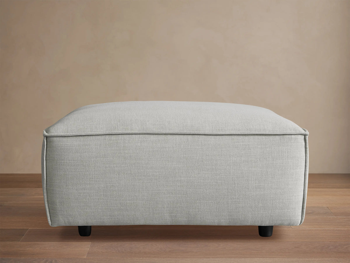 Light gray Coburn 38" ottoman, upholstered with subtle piping, on a wooden floor against a light brown wall.