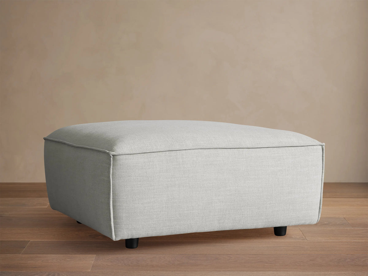 Light gray Coburn 38" ottoman, upholstered with subtle piping, on a wooden floor against a light brown wall.