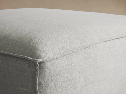 Coburn 38" light gray ottoman, rectangular with soft edges, prominent piping, and small dark feet on a wood floor.