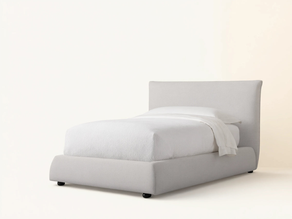 Rigby Twin Bed with light greige upholstered frame, tall headboard, and curved footboard, made up with white bedding.