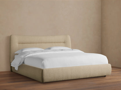 Lille Bed in fawn textured fabric with a curved upholstered headboard featuring horizontal lines, white bedding, on a wood floor.