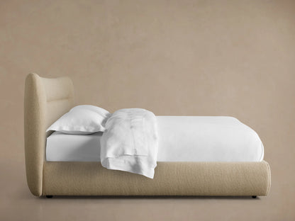 Side view of a Lille Bed featuring a fawn upholstered frame, curved headboard, and white bedding.